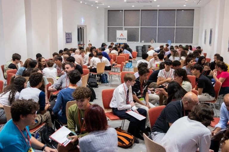 Many small groups of seated students in a large hall, interacting with their mentors.