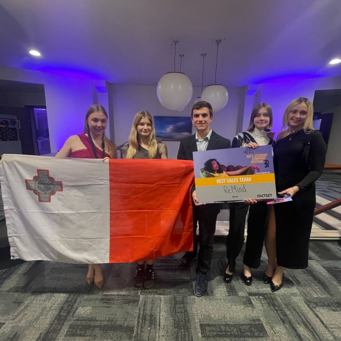 Team of JA Malta Company Programme students, standing proudly in a large carpeted hall, with the Maltese flag, holding an award