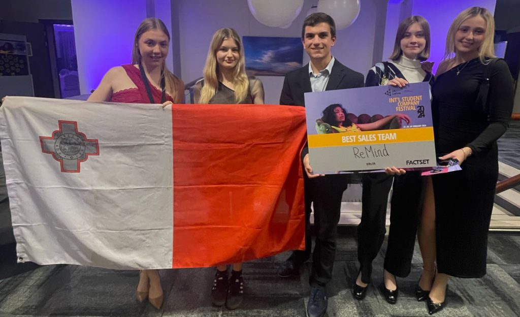 Team of JA Malta Company Programme students, standing proudly in a large carpeted hall, with the Maltese flag, holding an award