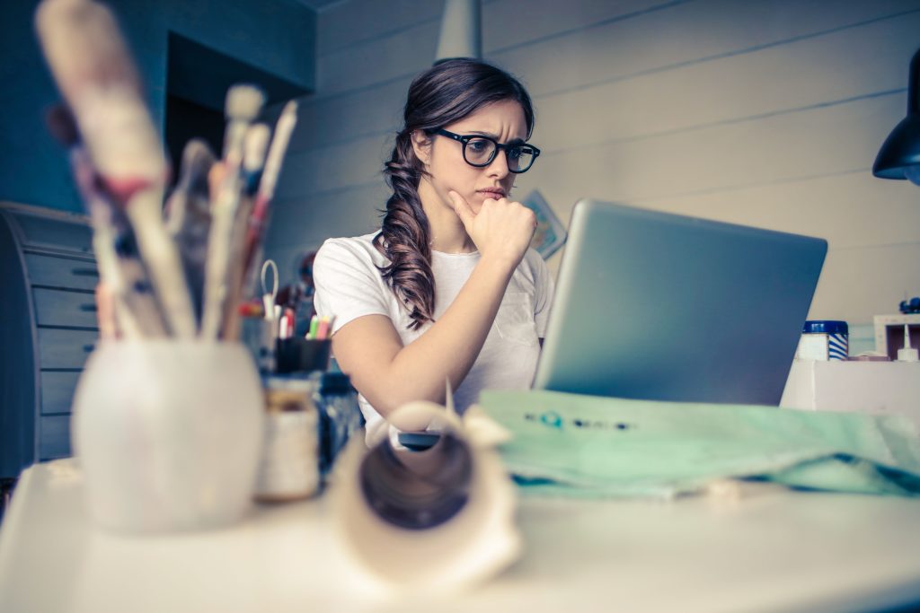 Young woman with glasses sitting at a desk looking at a laptop screen with a thoughtful expression