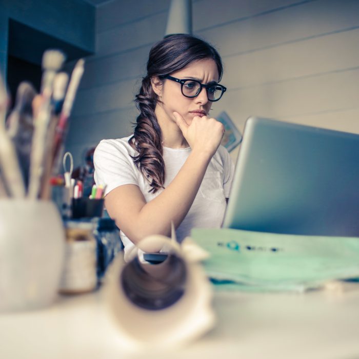 Young woman with glasses sitting at a desk looking at a laptop screen with a thoughtful expression