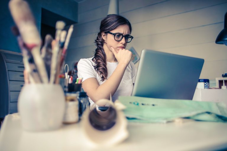 Young woman with glasses sitting at a desk looking at a laptop screen with a thoughtful expression